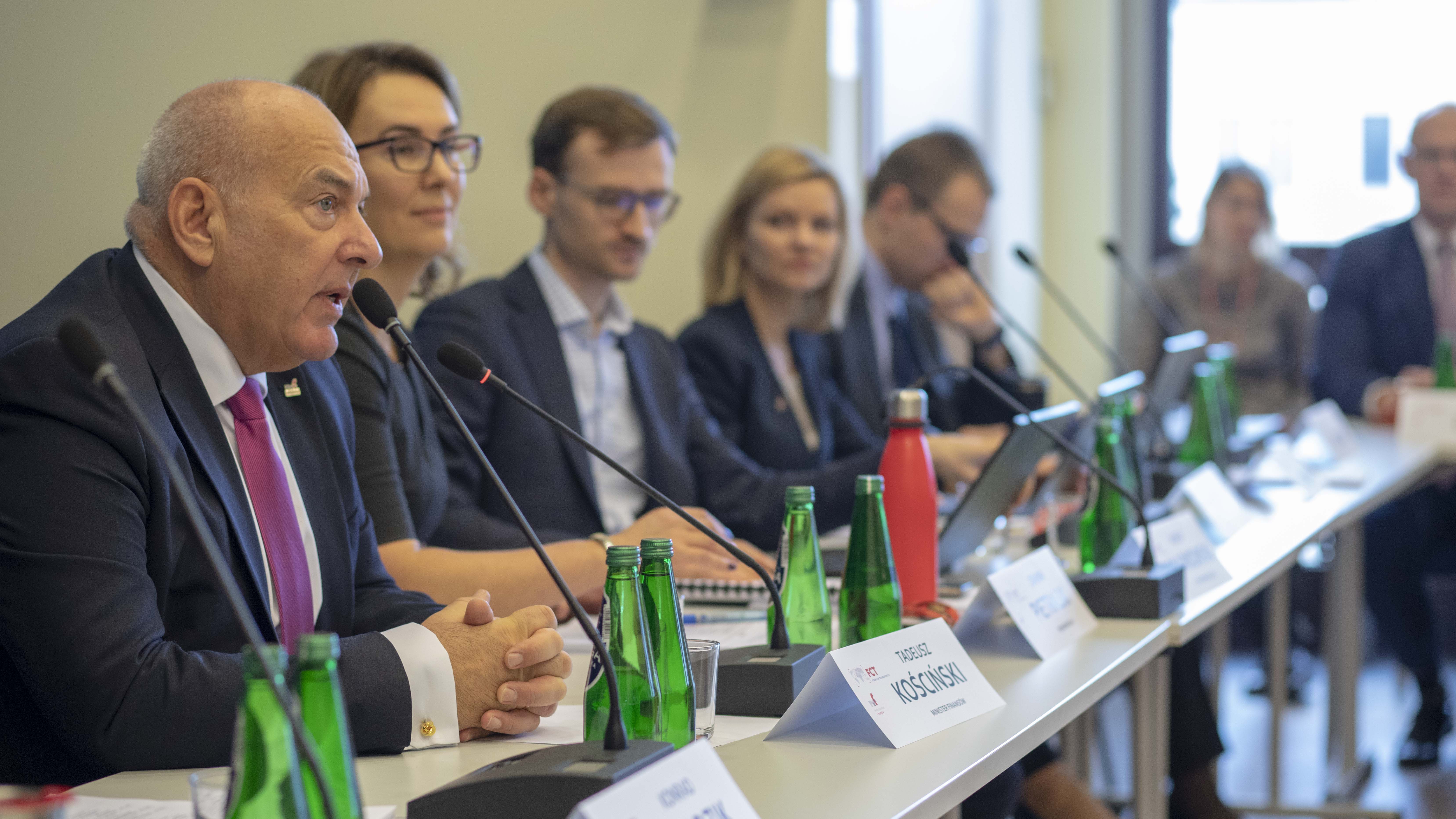 Rząd ludzi siedzących za stołem konferencyjnym, na stole mikrofony, tabliczki z nazwiskami oraz butelki wody. Pierwszy od lewej Minister Finansów - Tadeusz Kościński